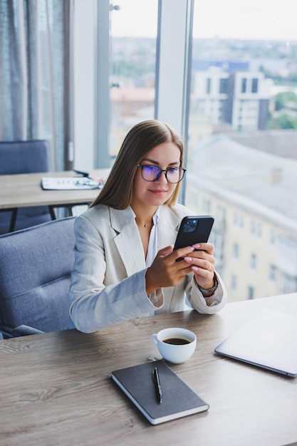 Mulher blogueira com celular digital sorrindo durante o intervalo para o café no interior do café mulher europeia feliz com smartphone e bebida de cafeína posando