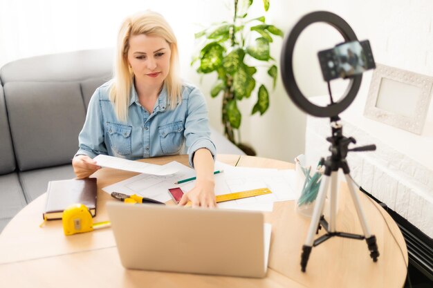 mulher blogueira gravando vídeo no smartphone e se iluminando com lâmpada de anel na mesa na sala de luz.