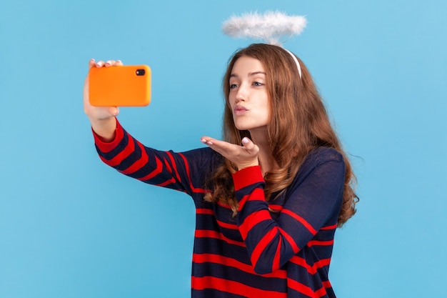 Mulher blogueira vestindo suéter listrado estilo casual e nimb sobre a cabeça transmitindo transmissão ao vivo enviando beijos aéreos para assinantes Estúdio interno isolado em fundo azul