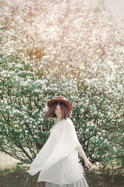 Mulher boho feliz no chapéu se divertindo na luz ensolarada perto da árvore branca florescendo no parque primavera Menina elegante hipster curtindo a primavera e dançando Momento atmosférico de felicidade