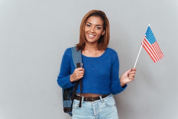 Mulher bonita africana sorridente em suéter e jeans com mochila e segurando a bandeira dos EUA. Fundo cinza isolado
