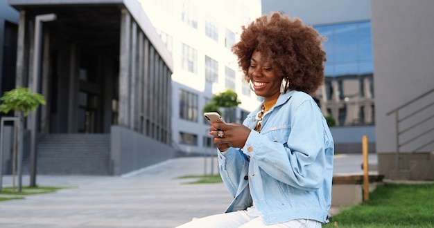 Mulher bonita afro-americana tocando e rolando no celular e sentado no banco do lado de fora. mensagens femininas atraentes elegantes no smartphone. conversa por telefone.