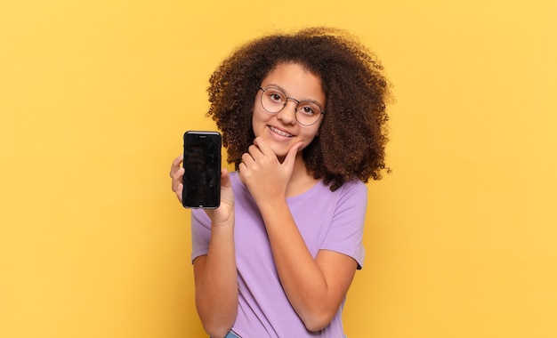 Mulher bonita afro sorrindo com uma expressão feliz e confiante com a mão no queixo, pensando e olhando para o lado e segurando uma cela