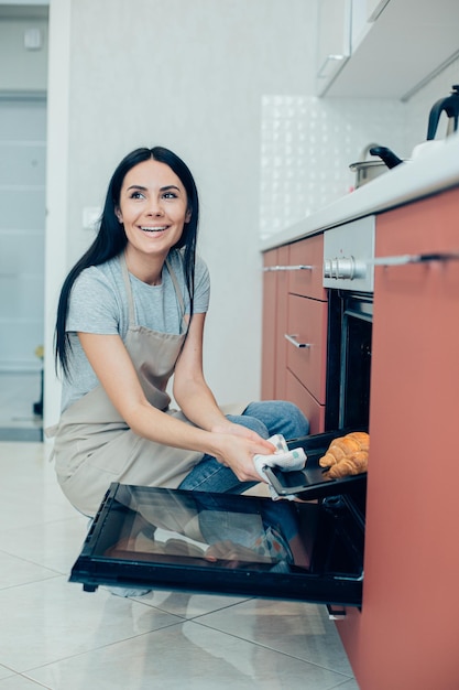 Mulher bonita alegre vestindo um avental e olhando para longe enquanto tira uma assadeira do forno