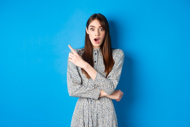 Mulher bonita animada em vestido apontando para canto superior esquerdo, olha com descrença e espanto, conferindo promo bacana, azul.