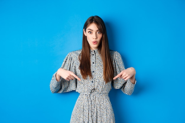 Mulher bonita animada no vestido diz uau, apontando os dedos para baixo e parece divertida, verificando a promoção, parada no azul.