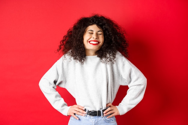 Mulher bonita animada rindo e sorrindo despreocupada, curtindo a vida, em pé de moletom contra o fundo vermelho.