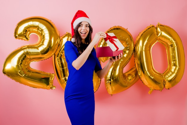 Mulher bonita animada usando chapéu de Papai Noel, abrindo a caixa de presente na frente de balões de ano novo 2020 isolados sobre rosa