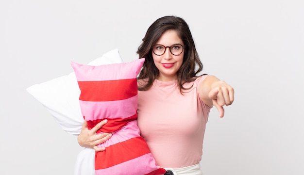 Mulher bonita apontando para a câmera escolhendo você de pijama e segurando um travesseiro