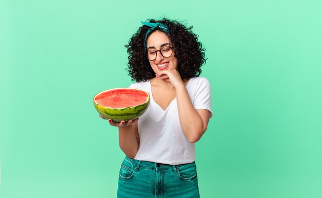 Mulher bonita árabe sorrindo alegremente e sonhando ou duvidando e segurando uma melancia. conceito de verão