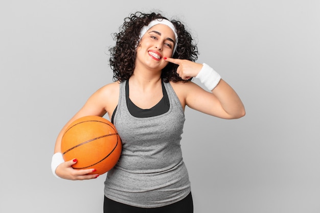 Mulher bonita árabe sorrindo com confiança, apontando para o próprio sorriso largo e segurando uma bola de basquete. conceito de esporte