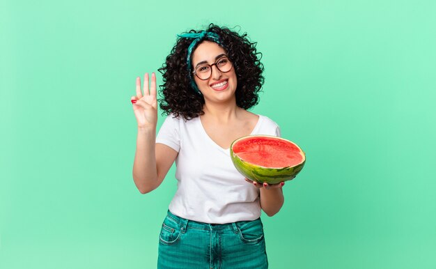 Mulher bonita árabe sorrindo e parecendo amigável, mostrando o número três e segurando uma melancia. conceito de verão