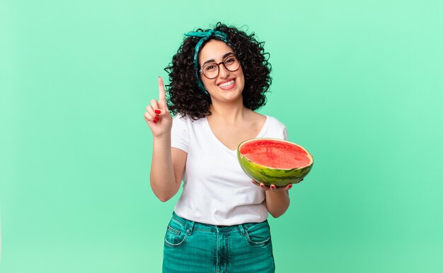 Mulher bonita árabe sorrindo e parecendo amigável, mostrando o número um e segurando uma melancia. conceito de verão