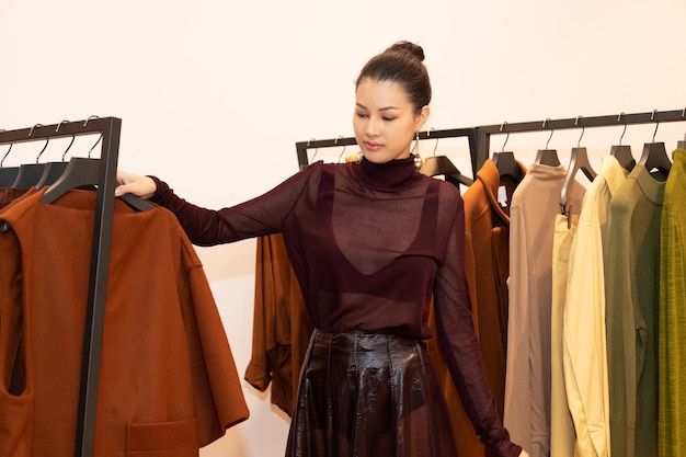 Mulher bonita asiática em vestido seleciona nova coleção em estante de roupas laranja verde tom terra em loja de moda de varejo que acaba de abrir novidades de marca para o outono de inverno como estilo minimalista