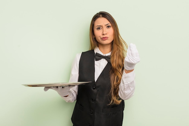 Mulher bonita asiática fazendo gesto de capice ou dinheiro dizendo para você pagar o conceito de garçom e bandeja