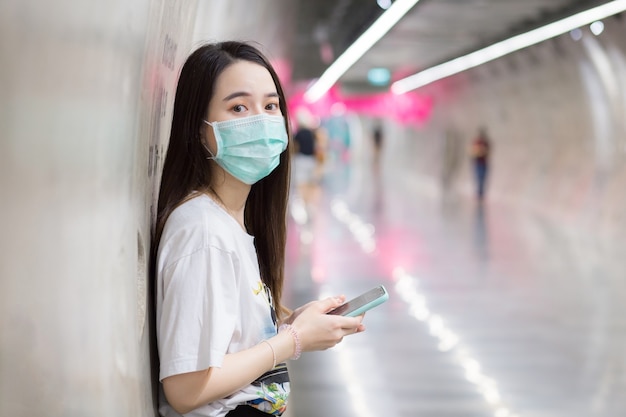 Mulher bonita asiática jovem está no túnel do metrô e usa máscara facial como novo estilo de vida normal e de saúde, enquanto segura o smartphone na mão.