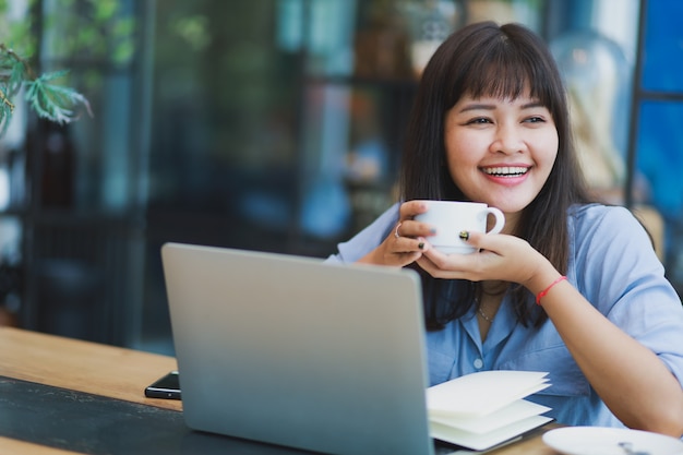 Mulher bonita asiática na camisa azul usando laptop e bebendo café