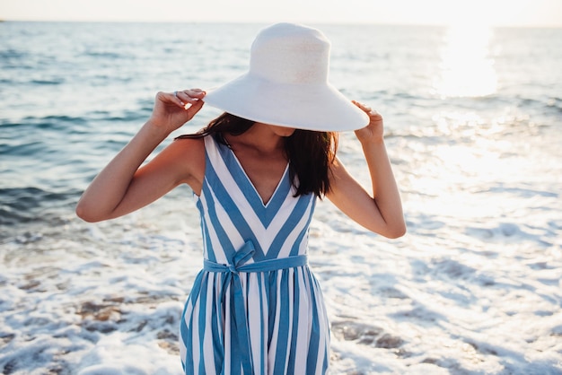 Mulher bonita atraente com chapéu branco na cabeça no fundo do pôr do sol ou nascer do sol do mar Ideia de capa Mulher sexy no vestido esconder o rosto com chapéu Conceito de viagem Conceito de moda de estilo de vida