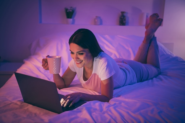 Mulher bonita, atraente e alegre deitada na cama usando um laptop