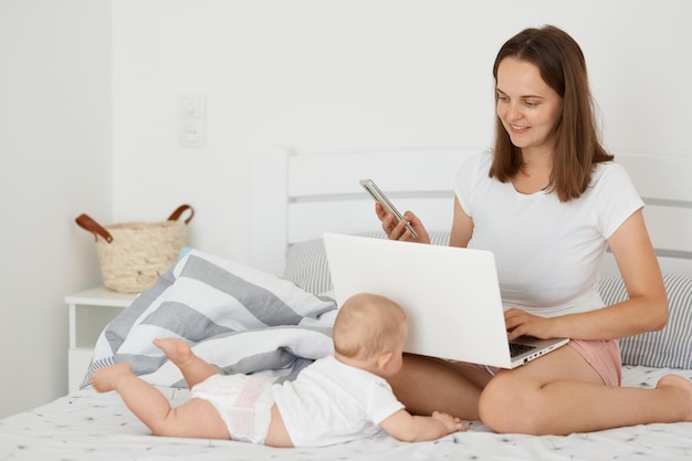 Mulher bonita bonita sentada na cama com sua filha infantil e trabalhando online segurando laptop e celular combina licença maternidade e trabalho online posando no quarto