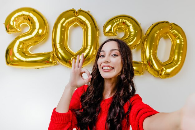 Mulher bonita camisola vermelha fazendo selfie engraçada na frente de 2020 balões de ano novo