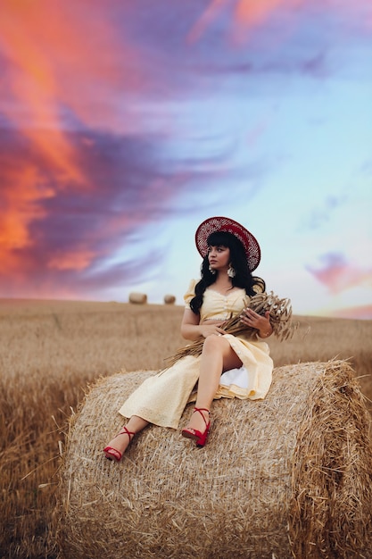 Foto mulher bonita caucasiana com cabelo preto comprido ondulado, vestido longo amarelo e chapéu vermelho, senta-se e relaxa