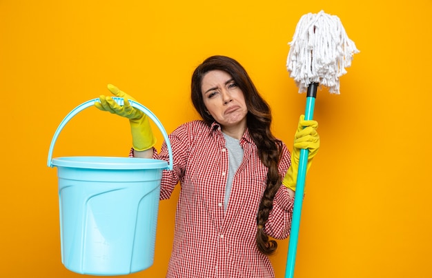 Mulher bonita caucasiana de limpeza descontente com luvas de borracha segurando um balde e um esfregão