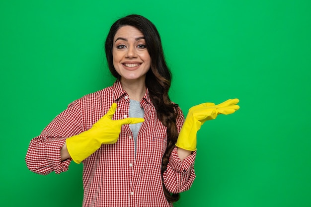 Mulher bonita caucasiana limpadora sorridente com luvas de borracha apontando para a mão vazia e olhando