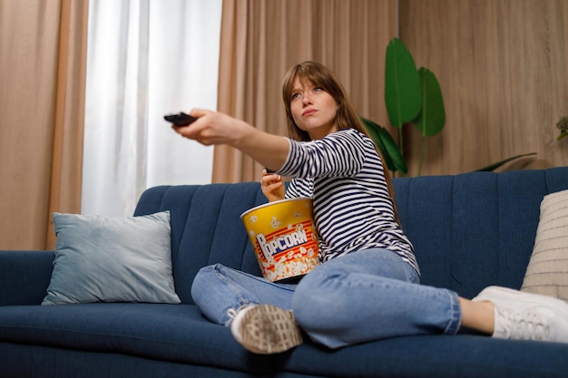 Mulher bonita caucasiana sentada no sofá comendo pipoca mudando de canal assistindo tv em casa