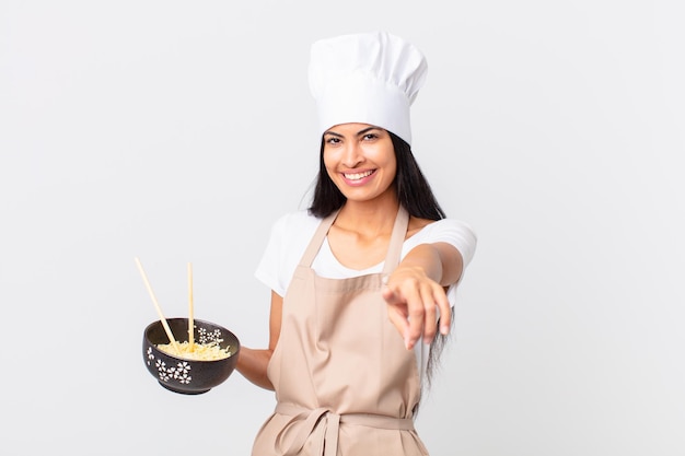 Mulher bonita chef hispânica apontando para a câmera escolhendo você e segurando uma tigela de macarrão