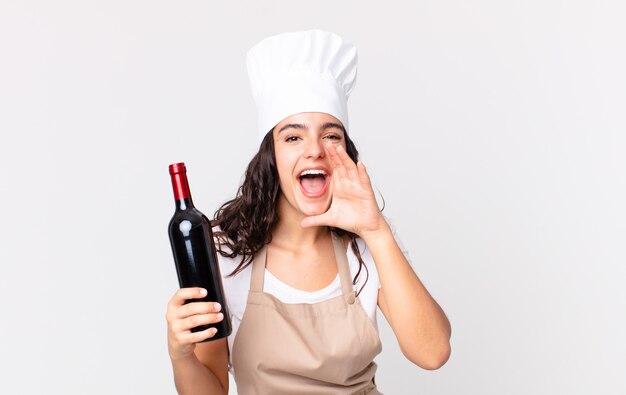 Mulher bonita chef hispânica se sentindo feliz, dando um grande grito com as mãos perto da boca e segurando uma garrafa de vinho