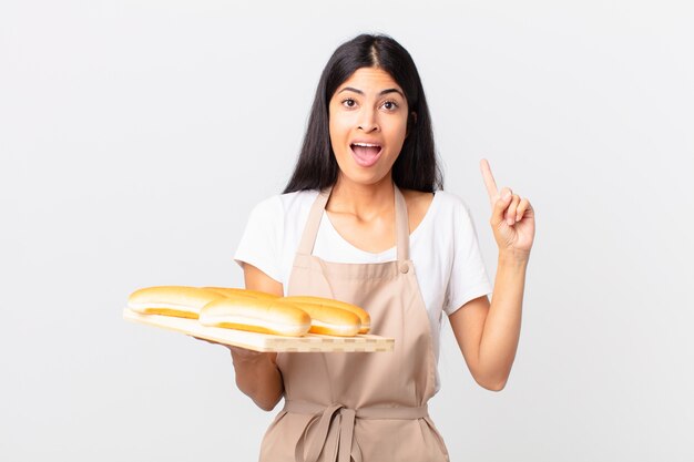 Mulher bonita chef hispânica se sentindo um gênio feliz e animado após ter uma ideia e segurando uma bandeja com pães