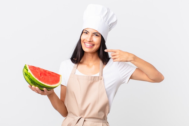 Mulher bonita chef hispânica sorrindo com confiança, apontando para o próprio sorriso largo e segurando uma melancia