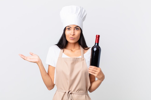 Mulher bonita chefe hispânica se sentindo perplexa e confusa, duvidando e segurando uma garrafa de vinho