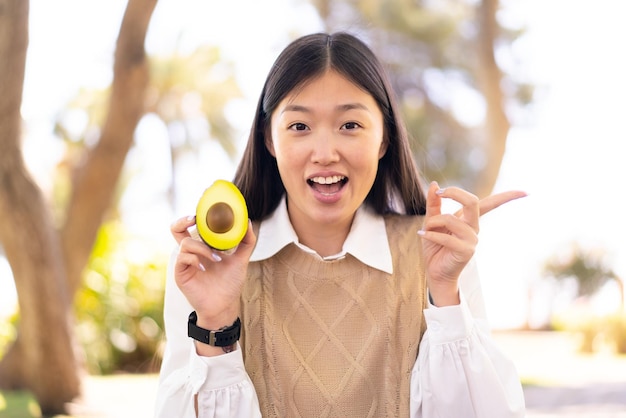 Mulher bonita chinesa segurando um abacate ao ar livre surpreso e apontando o dedo para o lado