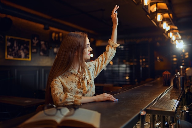 Mulher bonita com a mão levantada, sentado no balcão do bar. Uma pessoa feminina em um bar, emoções humanas, atividades de lazer, vida noturna