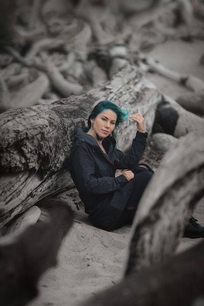 Mulher bonita com cabelo azul sentada na areia na praia cercada por árvores de toras