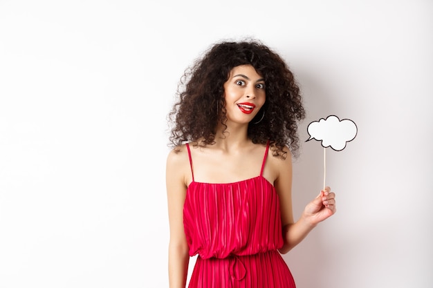 Mulher bonita com cabelo encaracolado, usando um vestido vermelho, segurando uma nuvem de comentários, diga algo, em pé sobre um fundo branco.