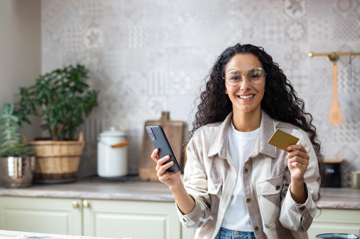 Foto mulher bonita com cabelo longo mantendo telefone e cartão de débito nas mãos em fundo de cozinha aconchegante dona de casa latina em óculos terminando compras on-line com entrega expressa espaço de cópia