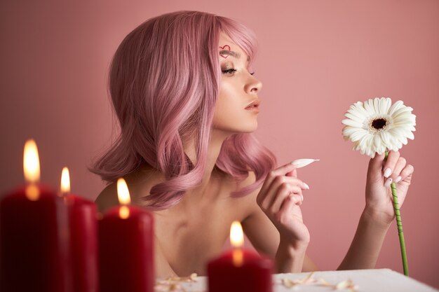 Mulher bonita com cabelo tingido de rosa, adivinhando uma flor na mão na mesa com velas.