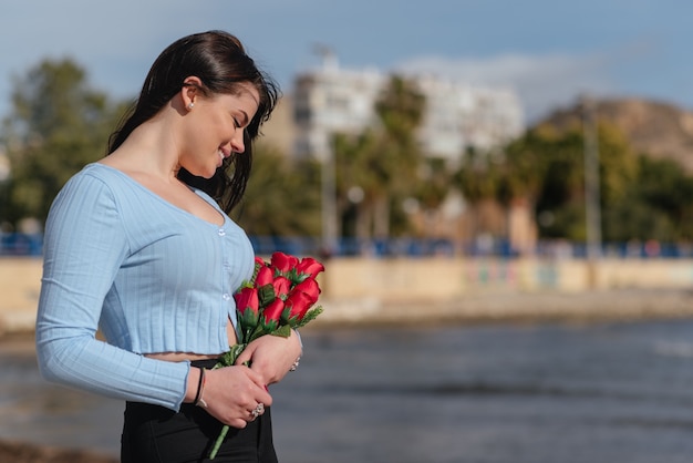 Mulher bonita com camiseta azul e calça preta segurando um buquê de rosas vermelhas ao ar livre no dia dos namorados