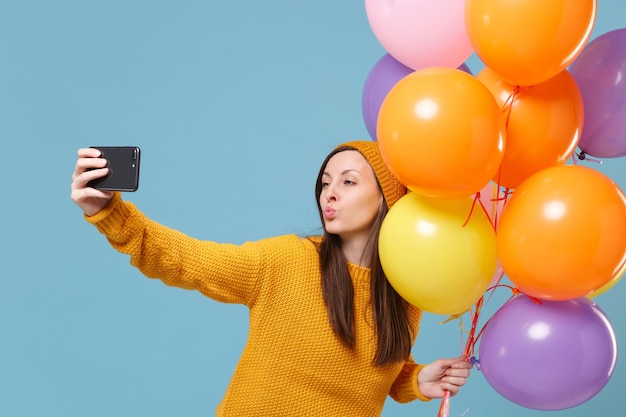 Mulher bonita com chapéu de suéter posando isolado sobre fundo azul. Festa de aniversário, conceito de emoções de pessoas. Mock up espaço de cópia. Celebrando segurar balões de ar fazendo selfie tiro no celular.