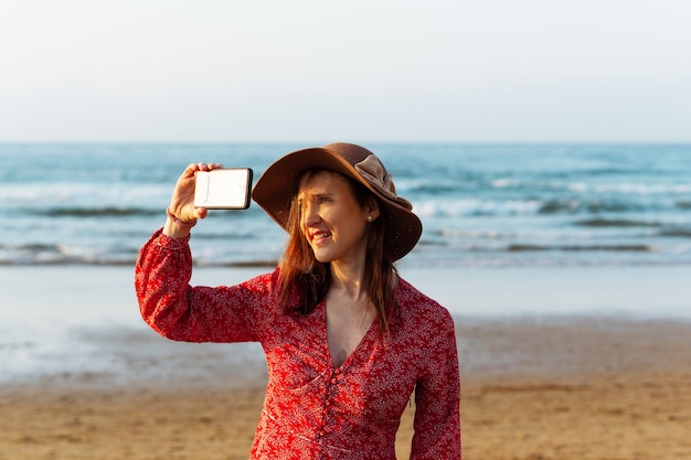 Mulher bonita com chapéu e vestido vermelho tirando uma selfie ou uma foto na primavera na praia na Espanha