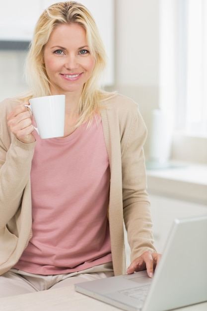 Mulher bonita com copo de café com laptop na cozinha