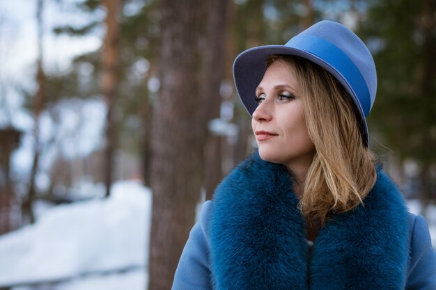Mulher bonita com maquiagem, com um casaco azul e um chapéu, posando na natureza na neve