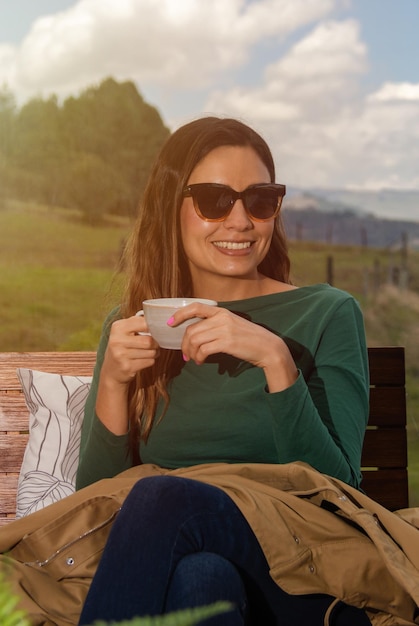 Mulher bonita com óculos de sol sorrindo tomando uma bebida quente com um fundo natural