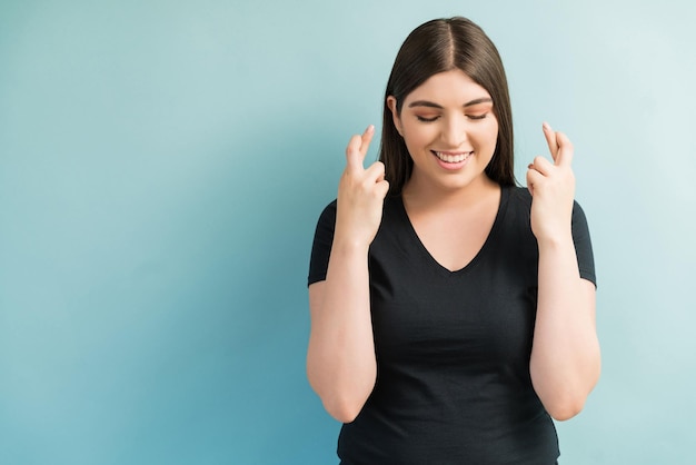 Mulher bonita com os olhos fechados e os dedos cruzados esperando boas notícias contra um fundo simples