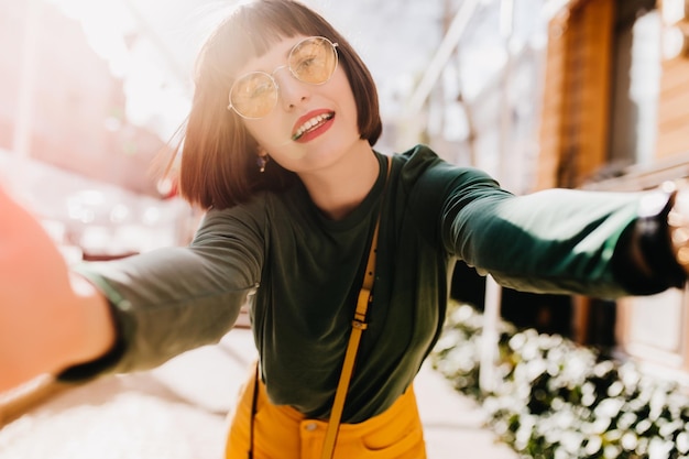 Mulher bonita com roupa elegante de primavera fazendo selfie Foto ao ar livre de modelo feminino espetacular com cabelo castanho curto tirando foto de si mesma no fundo da cidade