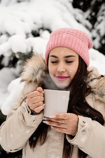 Mulher bonita com roupas quentes de inverno segurando uma xícara, bebendo chá quente ou café ao ar livre em dia de neve