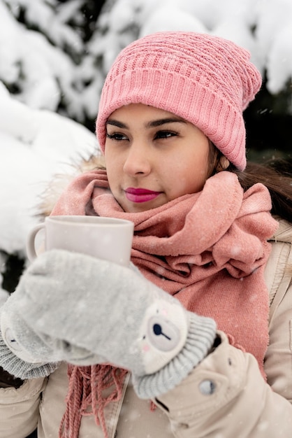 Mulher bonita com roupas quentes de inverno segurando uma xícara, bebendo chá quente ou café ao ar livre em dia de neve